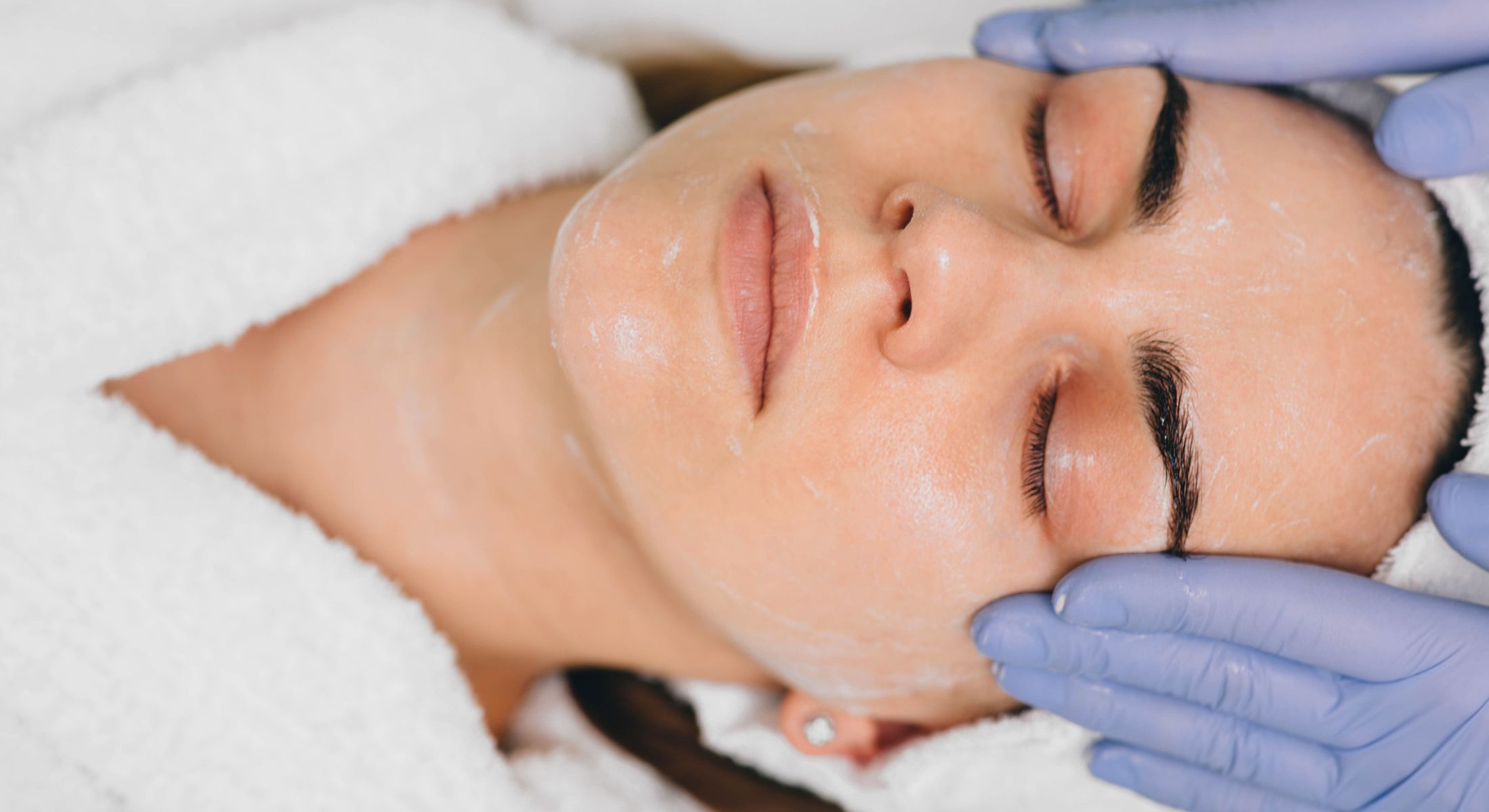 Facial treatment with gloved hands applying cream.